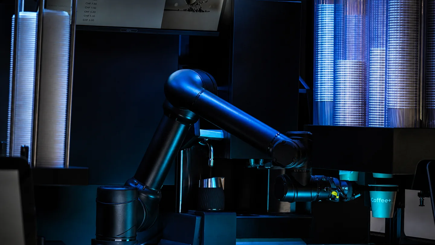 Der Coffee+ Roboterarm zwischen Becherstapeln und Milchkanne.